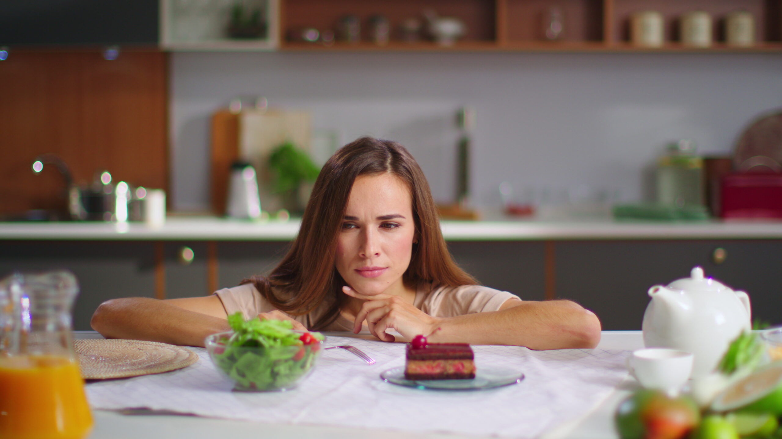 Portrait of smiling woman making decision between salad and cake on kitchen. Girl looking at cake and salad on table. Woman making choice between healthy or unhealthy food on kitchen