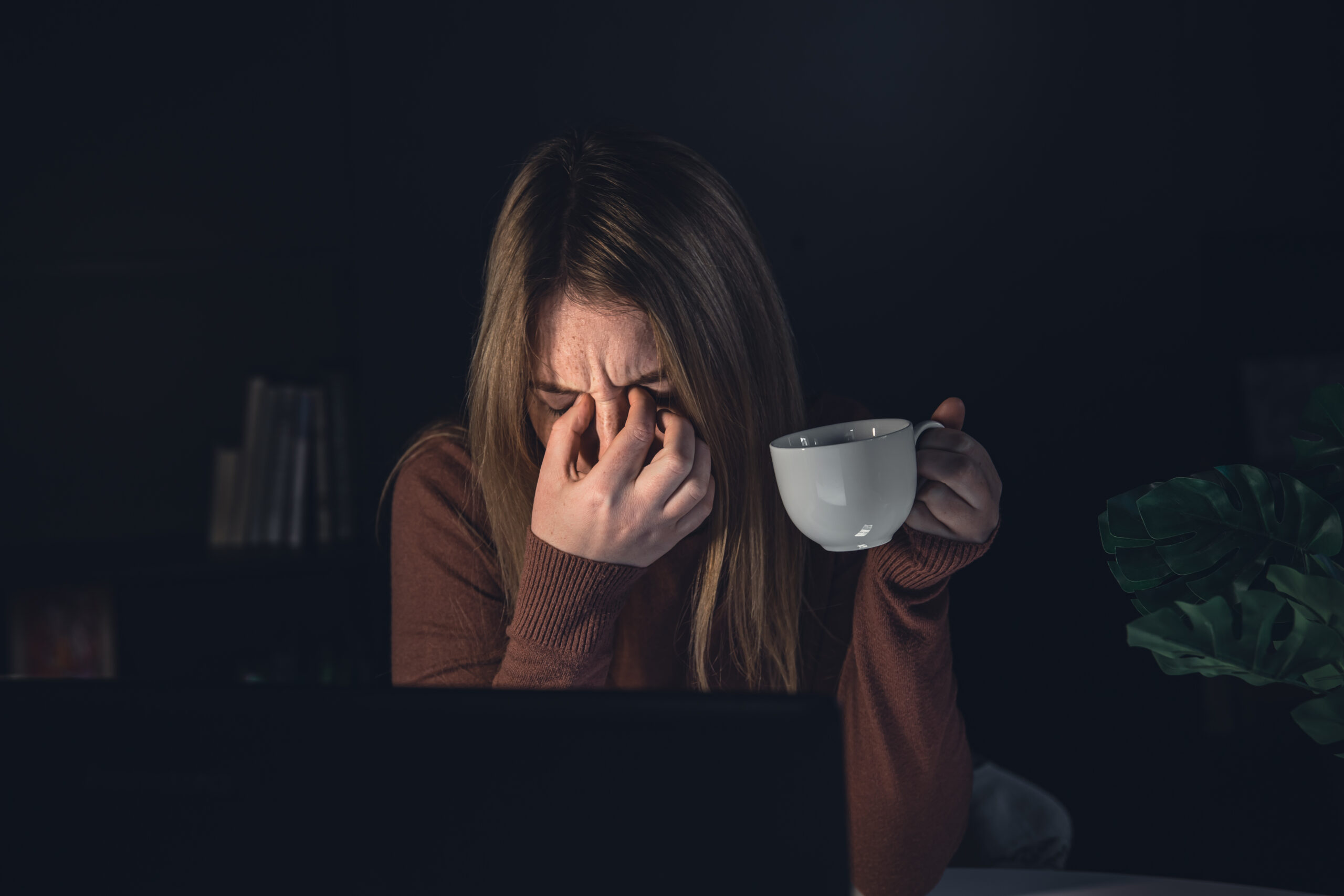 A young woman is working at a computer at night, holding a cup of coffee in her hands, wants to sleep.