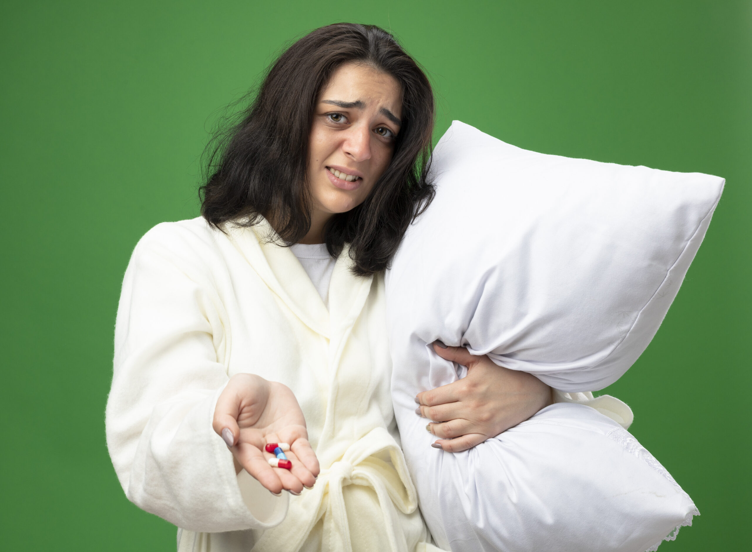 irritated young caucasian ill girl wearing robe holding pillow stretching out medical capsules towards camera looking at camera isolated on green background