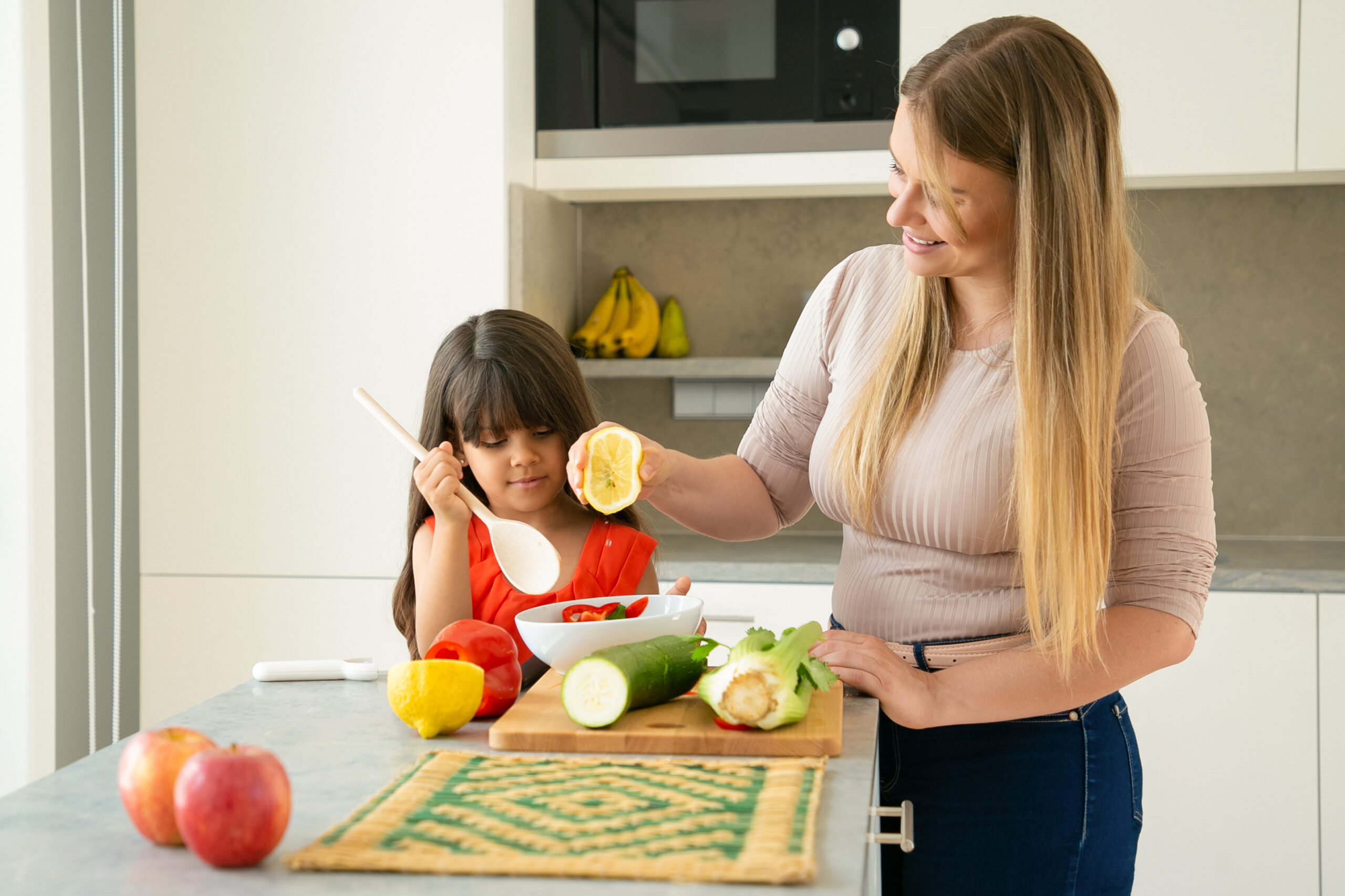 Mom and daughter cooking vegs for dinner at kitchen table. Girl and her mother dressing salad in bowl with half of lemon. Family cooking or healthy nutrition concept