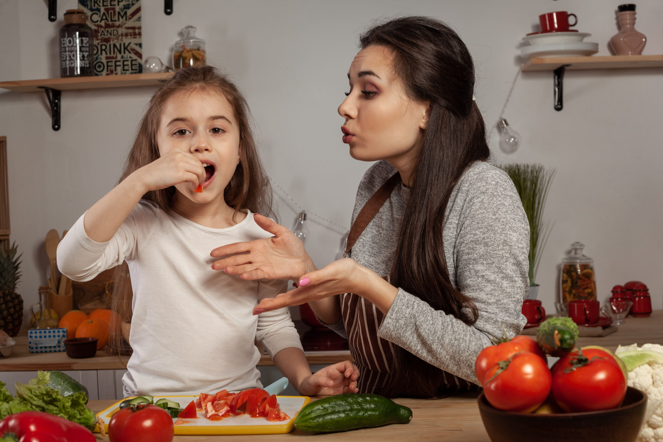 Happy loving family are cooking together. Beautiful mommy and her little girl are making a vegetable salad and tasting it at the kitchen, against a white wall with shelves and bulbs on it. Homemade food and little helper.
