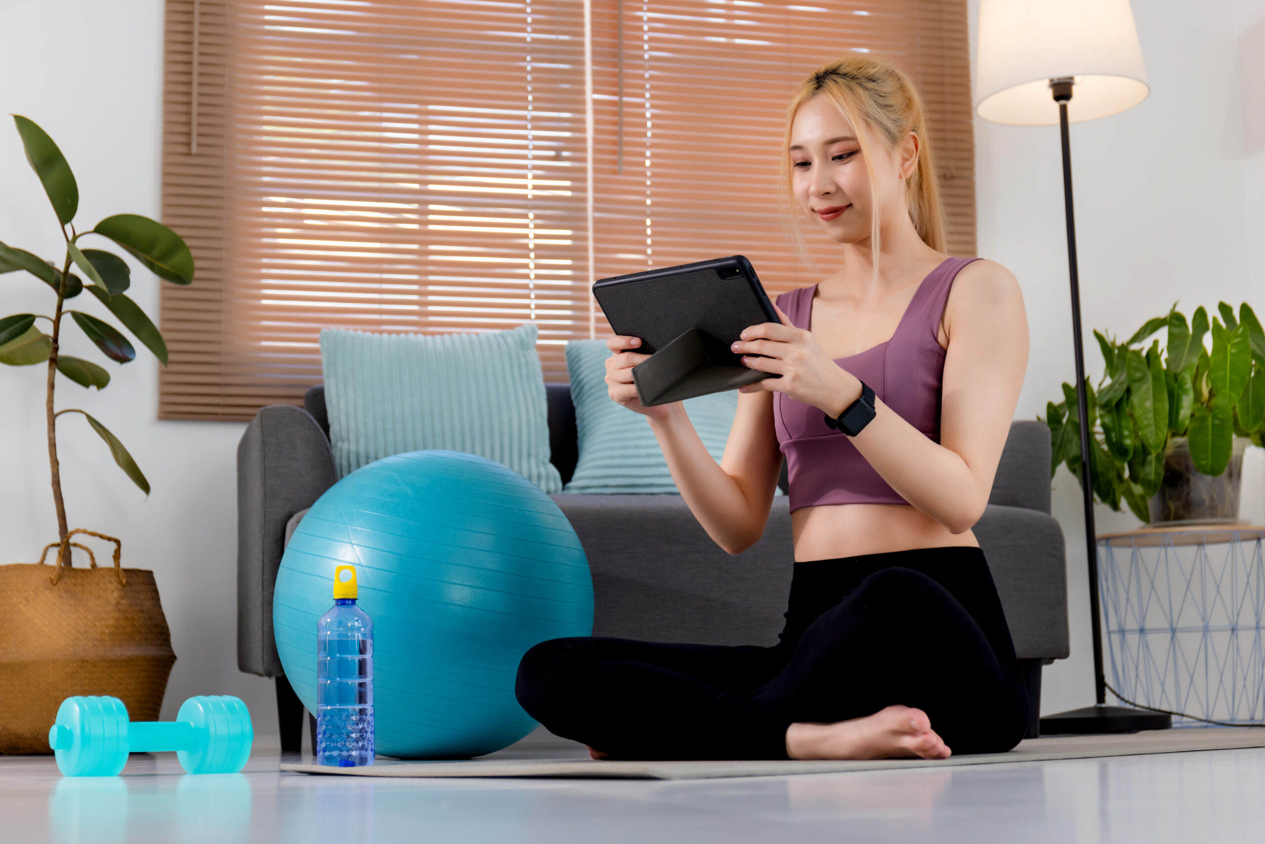 Pretty young Asian woman in sportswear sitting on exercise mat and watching training online videos on digital tablet.